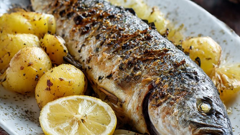 Pescado Lubina asado con patatas y rodajas de limón en un plato.