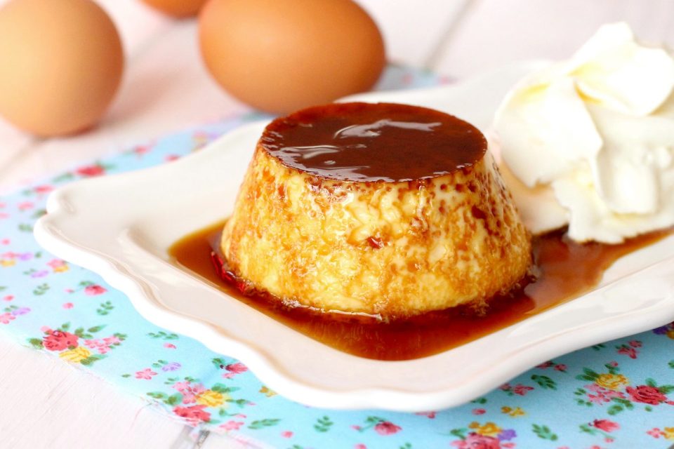 Flan con caramelo sobre plato, acompañado de nata y huevos al fondo.