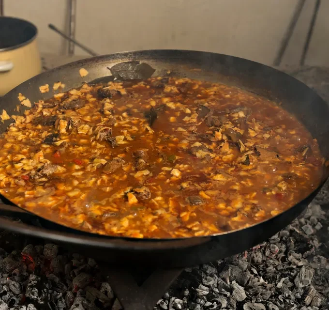 Paella cocinándose en una sartén grande sobre brasas, con carne y salsa espesa.